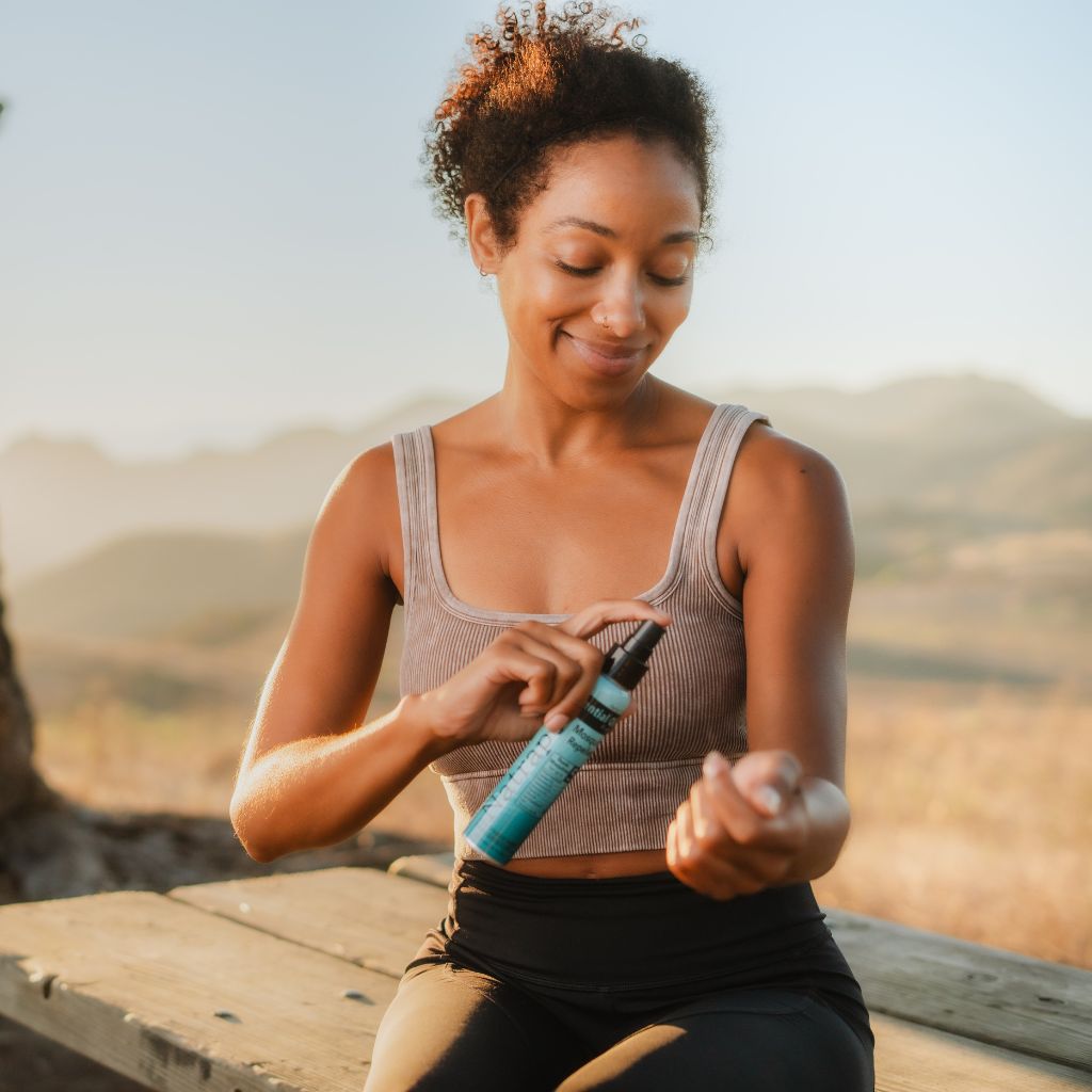 Woman using the Natrapel Essential Oil Insect Repellent spray on a wooden bench with a mountainous background